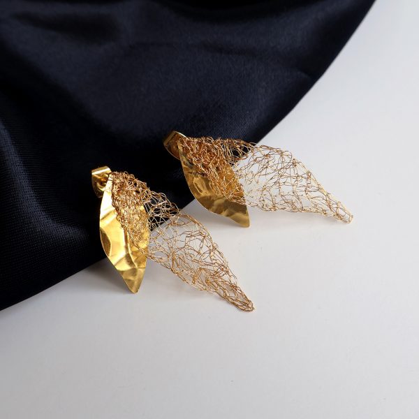 Les boucles d'oreilles Folÿs 2 sont des bijoux uniques brodés à la main par la marque Marie Archambaud. Elles reposent délicatement sur un tissu bleu marine, mettant en valeur l'éclat doré du métal. Elles sont composées de deux feuilles légères, une brodée au fil fin, et une en plaque de métal texturée, évoquant une nature poétique.